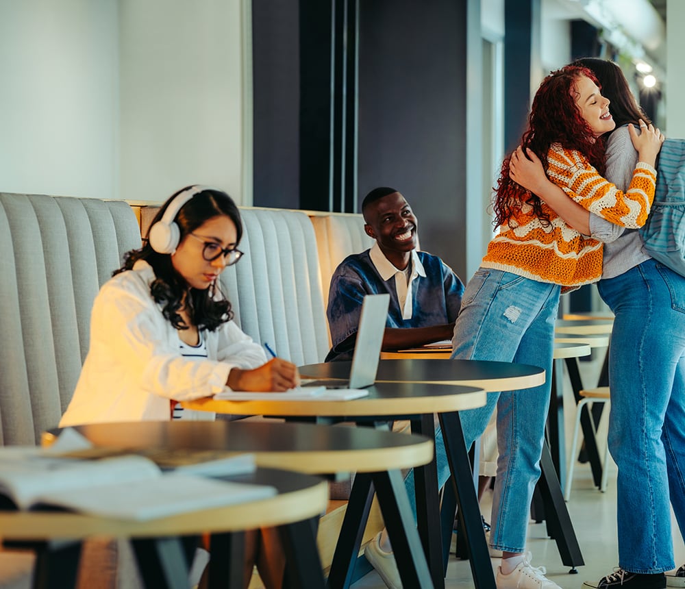 In a lively cafe setting, one person is working on a laptop, another is smiling warmly, and two are hugging near a table, capturing the essence of a vibrant campus community.