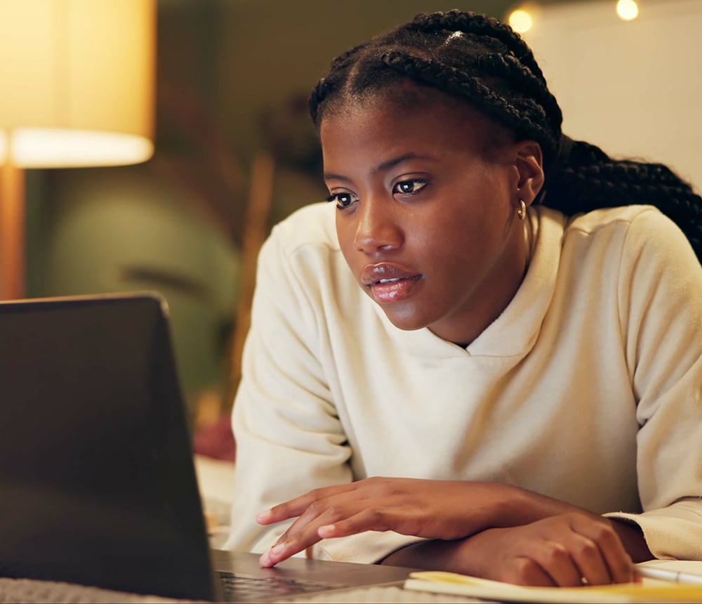 A person with braided hair is focused on a laptop screen in a warmly lit room, likely pondering over plans for the campus ministry to foster a sense of community.