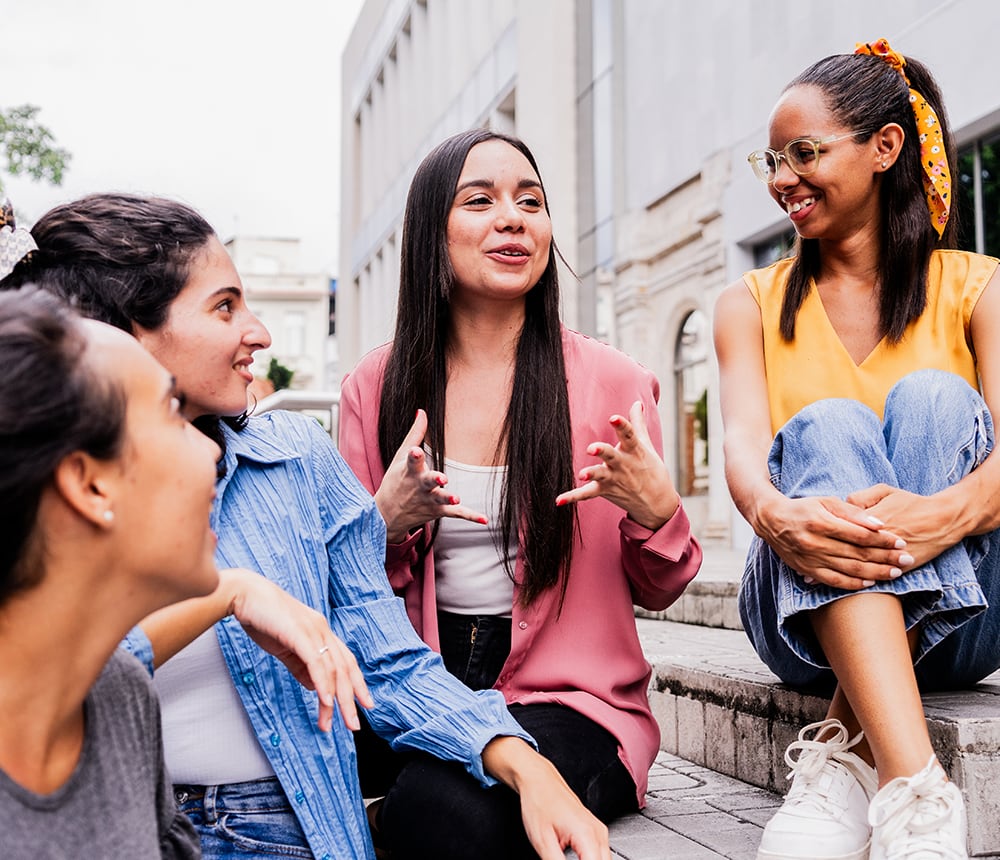 Four women, engaged in high school outreach, sit outdoors, smiling and chatting in a relaxed manner.