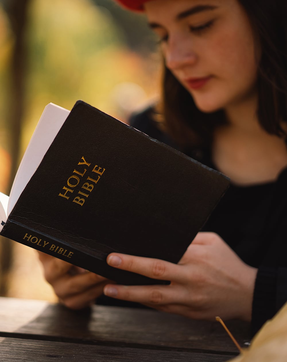 A student participating in a high school outreach program sits at a wooden table outdoors, thoughtfully reading a black Holy Bible.