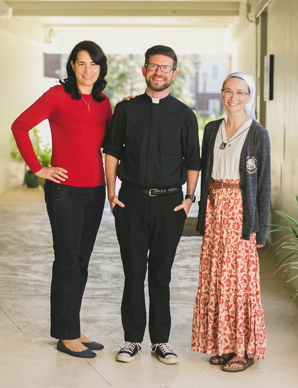 Three people stand in a warm corridor, beaming. The central figure, wearing a clerical collar, seems at home among friends.