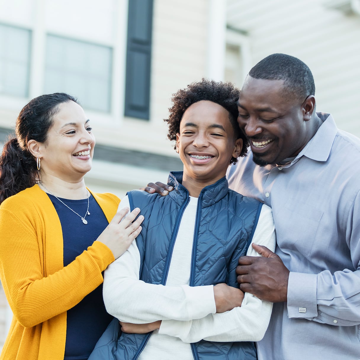 A smiling family of three stands together outside, with the parents happily embracing their child.