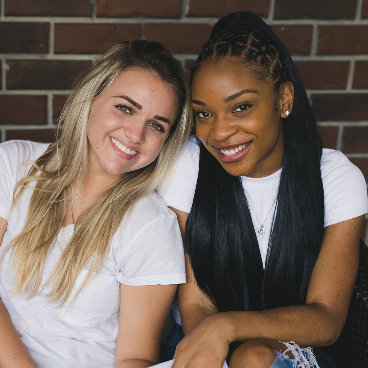 Two women smiling, sitting close together in front of a brick wall, both wearing white tops.